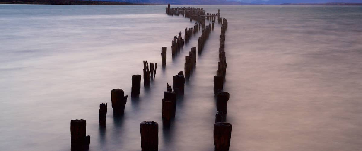 A long, straight wooden pier extending into a calm lake, surrounded by mountains and a cloudy sky.