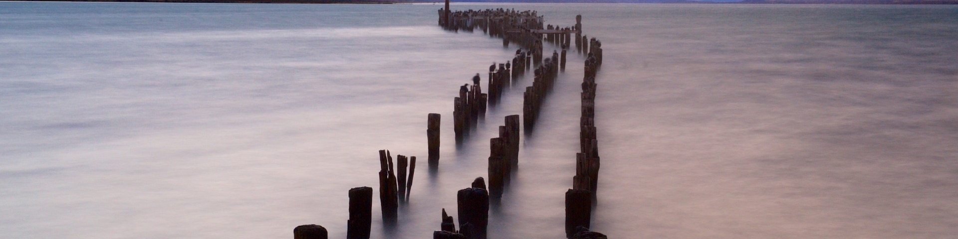 A long, straight wooden pier extending into a calm lake, surrounded by mountains and a cloudy sky.