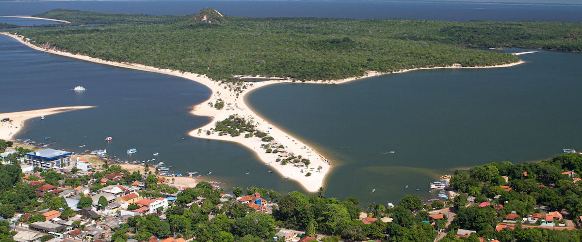 An aerial view of a coastal town with a beach, boats, and a forested peninsula.