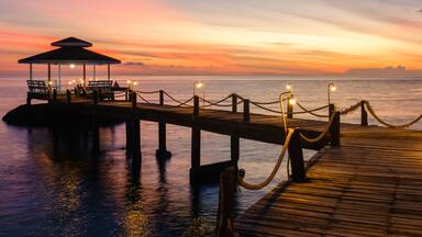 Experience the tranquil charm of Koh Kood Island in Thailand as the sun sets over the calm ocean waters. A wooden pier with lamps illuminates the peaceful scene, inviting relaxation.