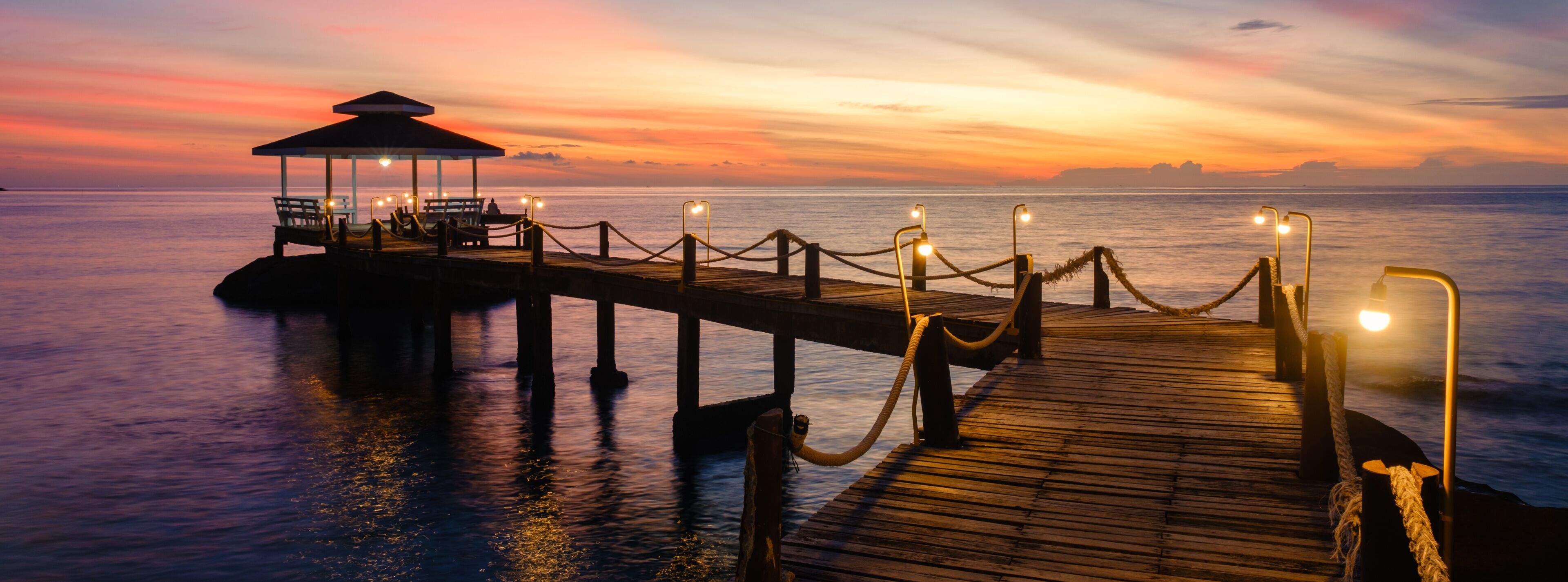 A wooden pier extending into calm waters at sunset, with a covered seating area and lights along the path.