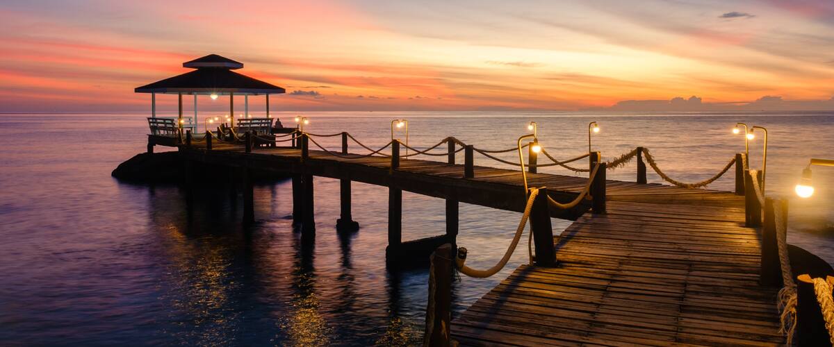 A wooden pier extending into calm waters at sunset, with a covered seating area and lights along the path.