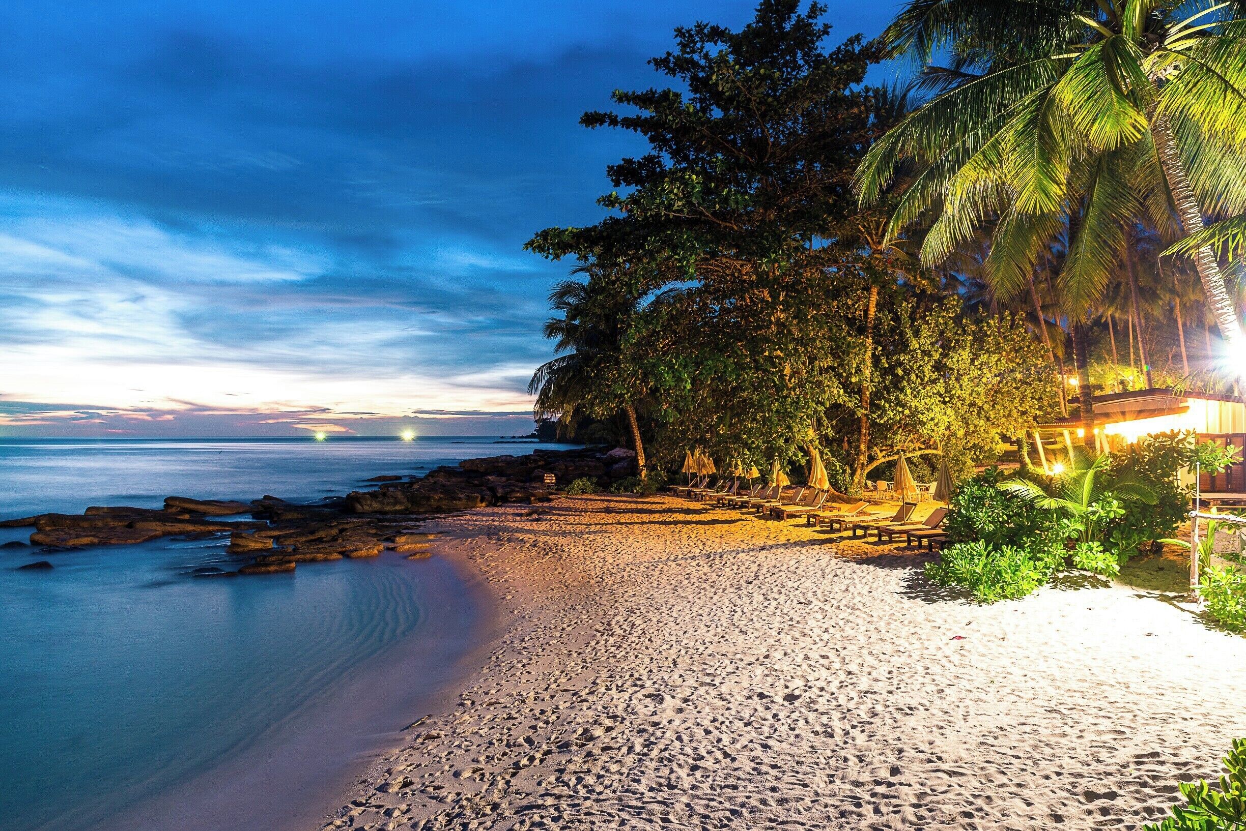 A beach with palm trees, a rocky shoreline, and a sunset.