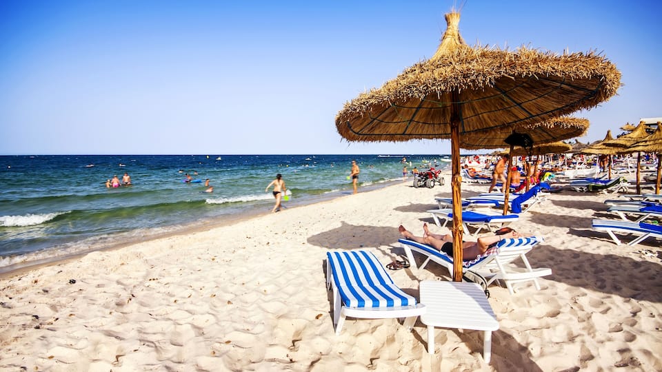 A beach with thatched umbrellas, sun loungers, and people in the water.