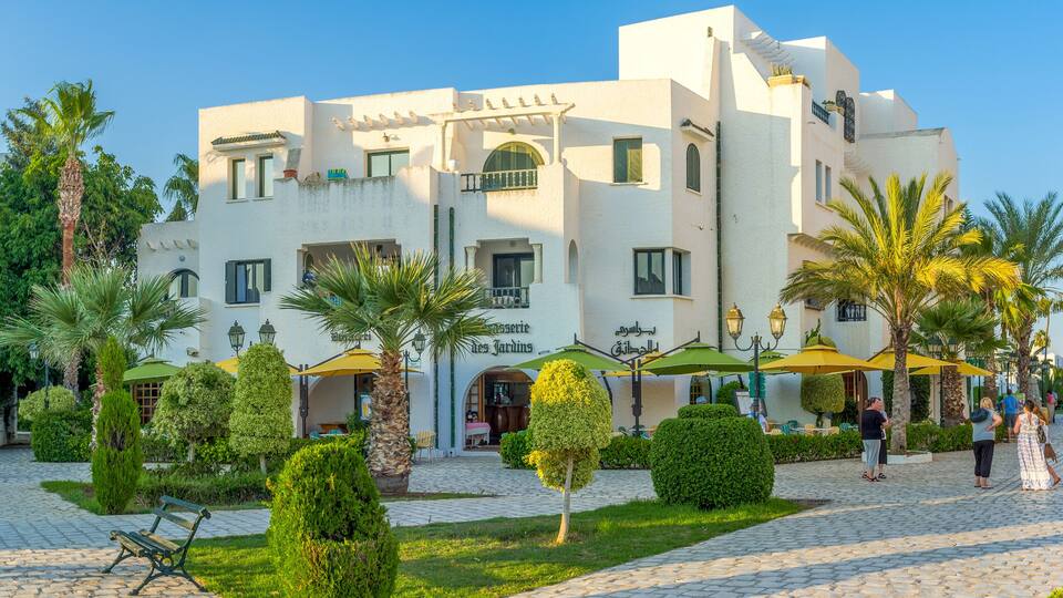 A white building with a café named "Les Jardins" and palm trees in the courtyard.