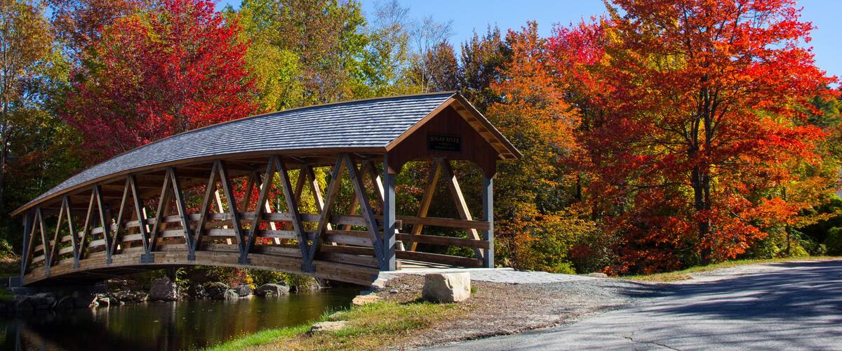 A wooden covered bridge over a calm river, surrounded by autumn foliage.