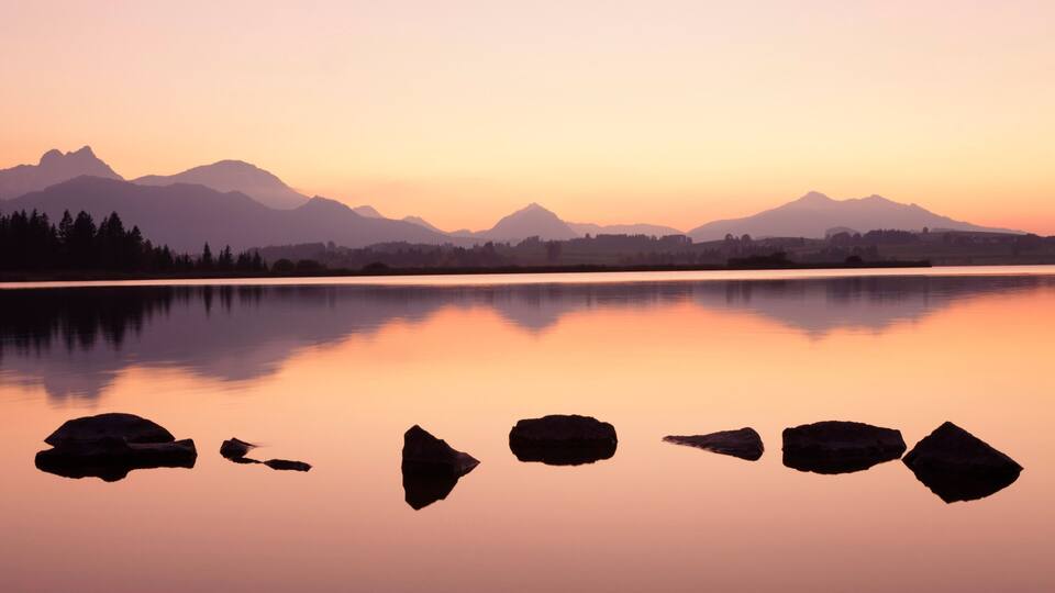 A calm lake with a mountain range in the background and several rocks in the foreground.