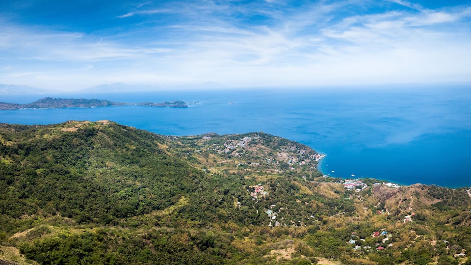 A coastal landscape with dense green hills, a clear blue sea, and scattered buildings.