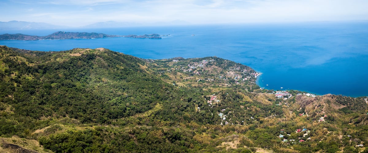 A coastal landscape with dense green hills, a clear blue sea, and scattered buildings.