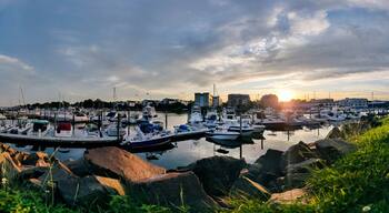 A marina with boats docked, surrounded by rocks and greenery, with buildings and a sunset in the background.