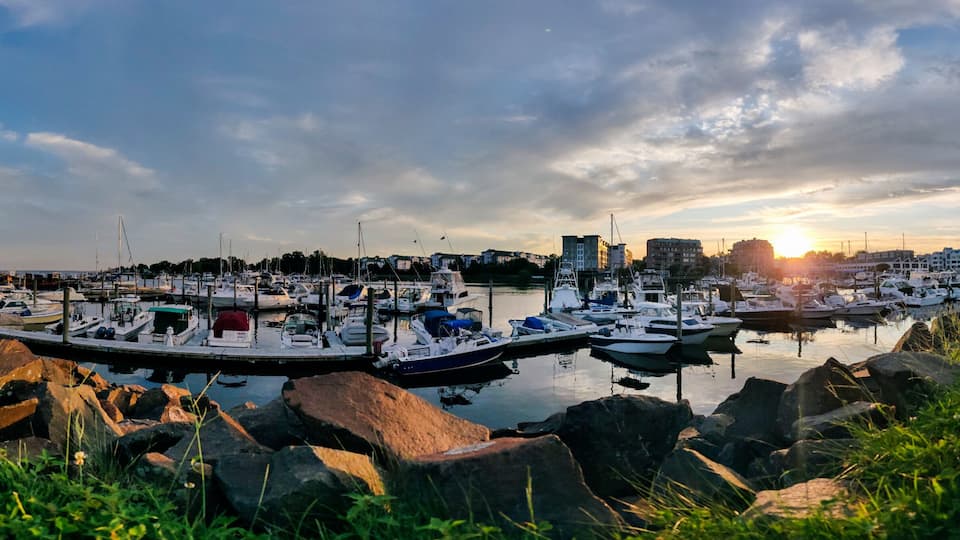En lysthavn med fortøjede både, omgivet af klipper og grønt, med bygninger og en solnedgang i baggrunden.