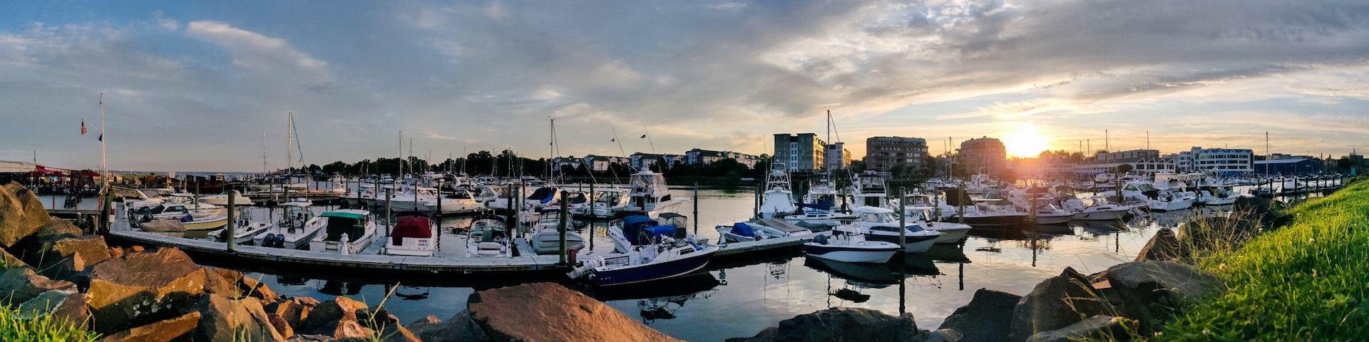 Un port de plaisance où sont amarrés des bateaux, entouré de rochers et de verdure, avec des bâtiments et un coucher de soleil en arrière-plan.