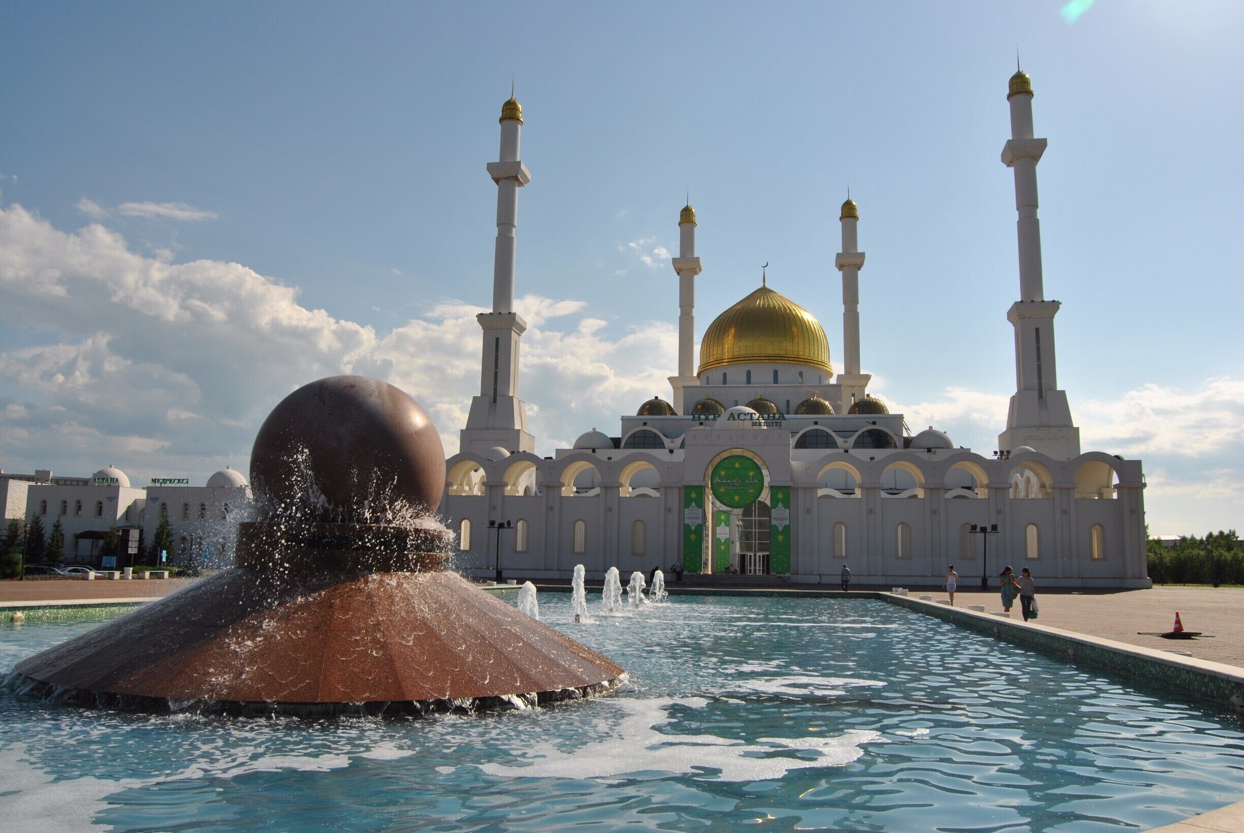 A mosque with a golden dome and four minarets, a large fountain in the foreground, and a clear blue sky.