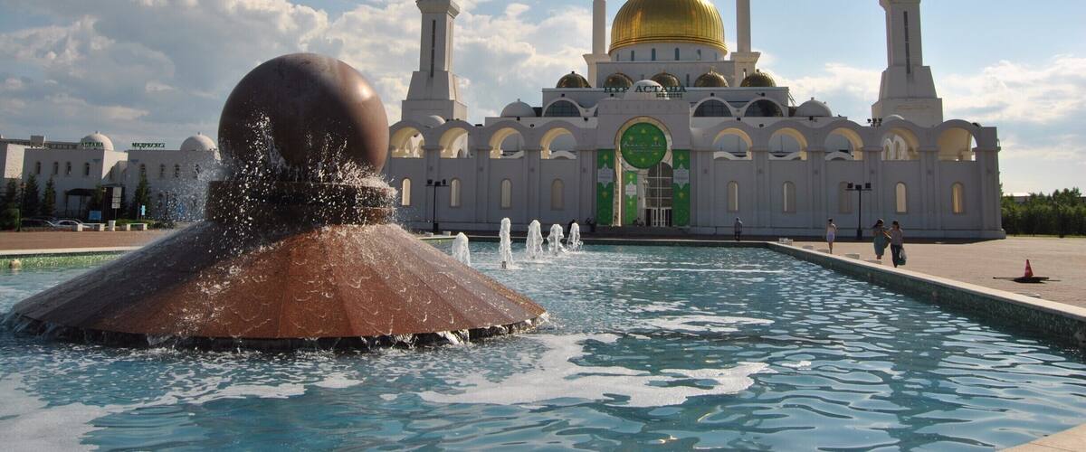 A mosque with a golden dome and four minarets, a large fountain in the foreground, and a clear blue sky.