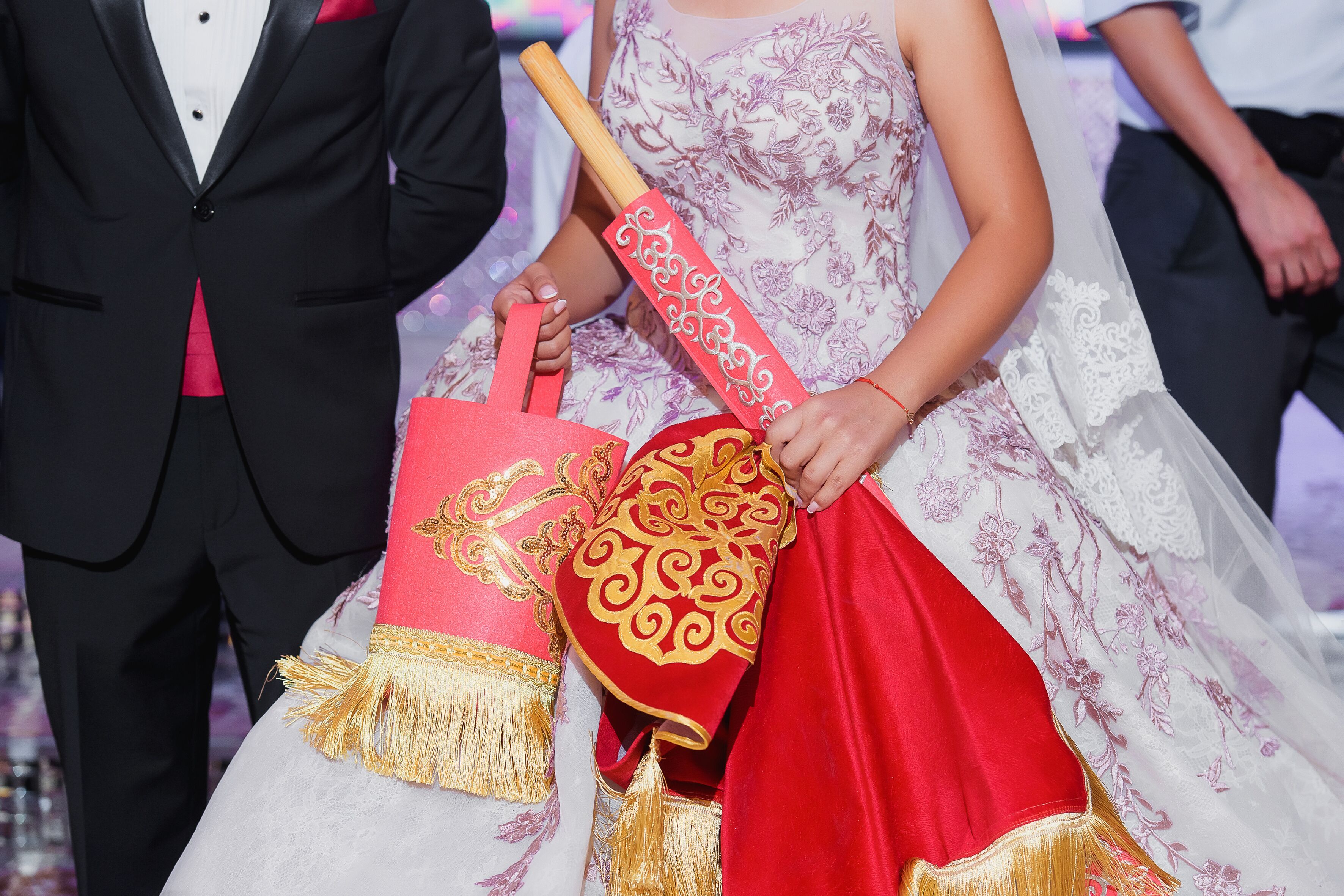 A person in a white dress with red and gold embroidery, holding a red and gold ceremonial fan.