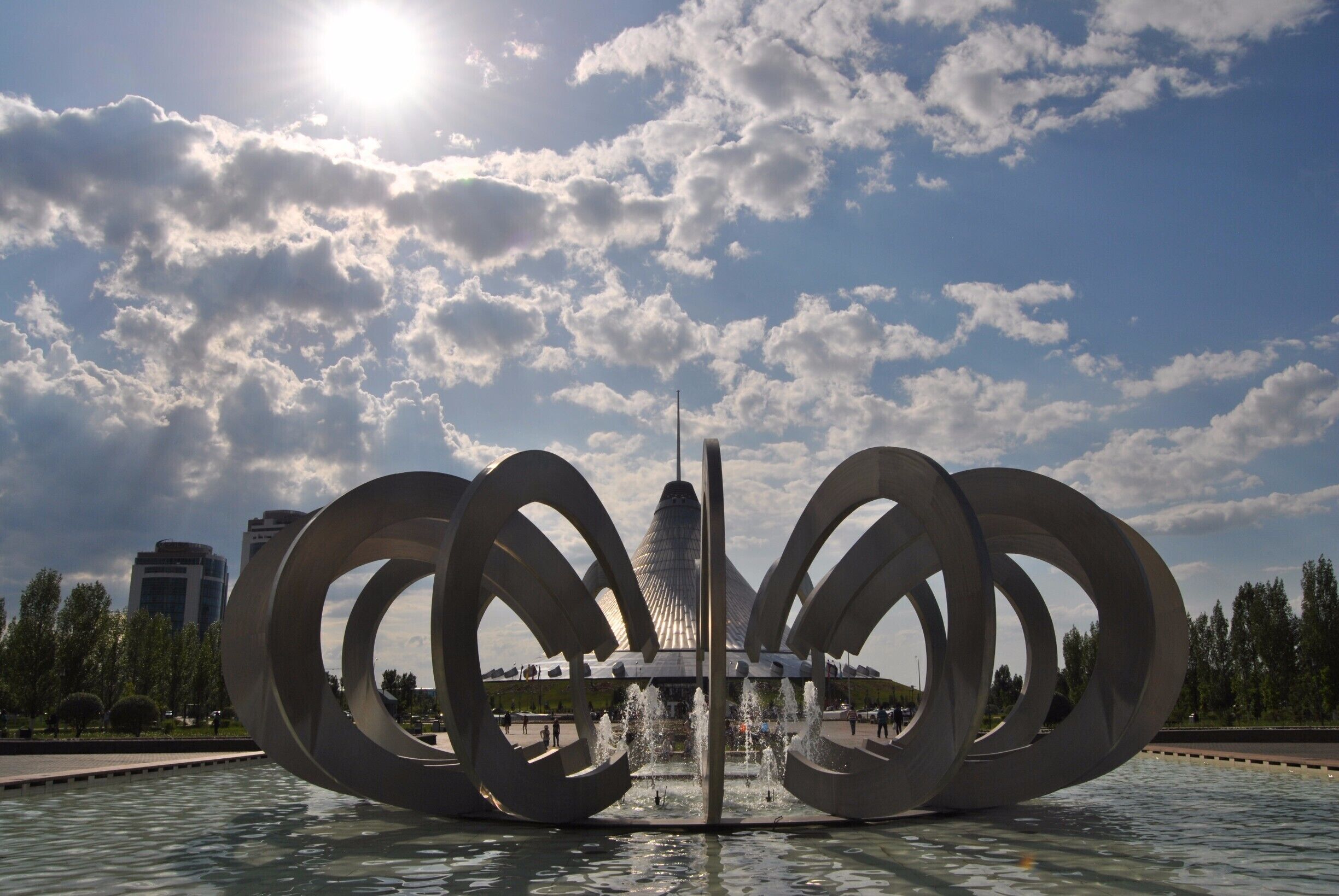 Una scultura moderna con anelli circolari in una piazza pubblica, con una fontana al centro, sotto un cielo parzialmente nuvoloso.