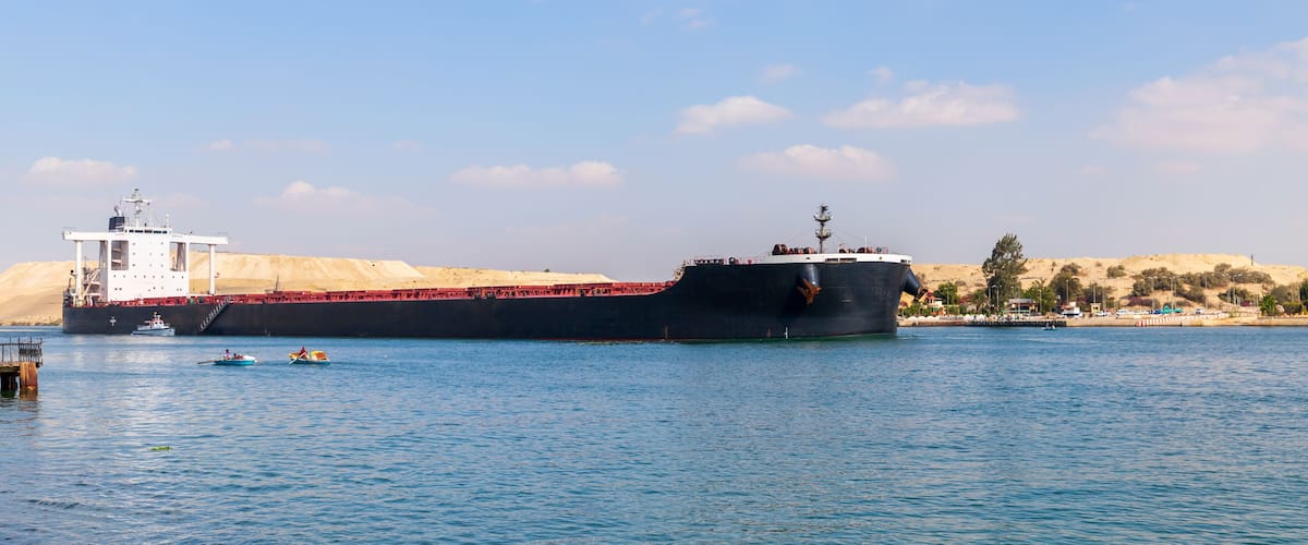 A large cargo ship docked at a port with a sandy landscape and a clear sky in the background.