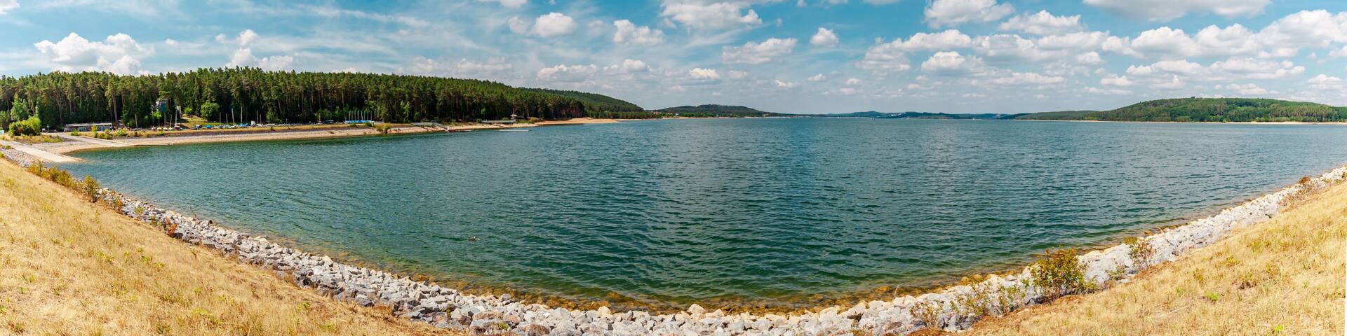 A calm lake with a rocky shoreline, surrounded by dense forests and distant hills under a partly cloudy sky.