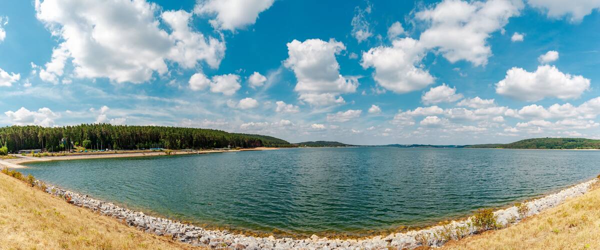 A calm lake with a rocky shoreline, surrounded by dense forests and distant hills under a partly cloudy sky.