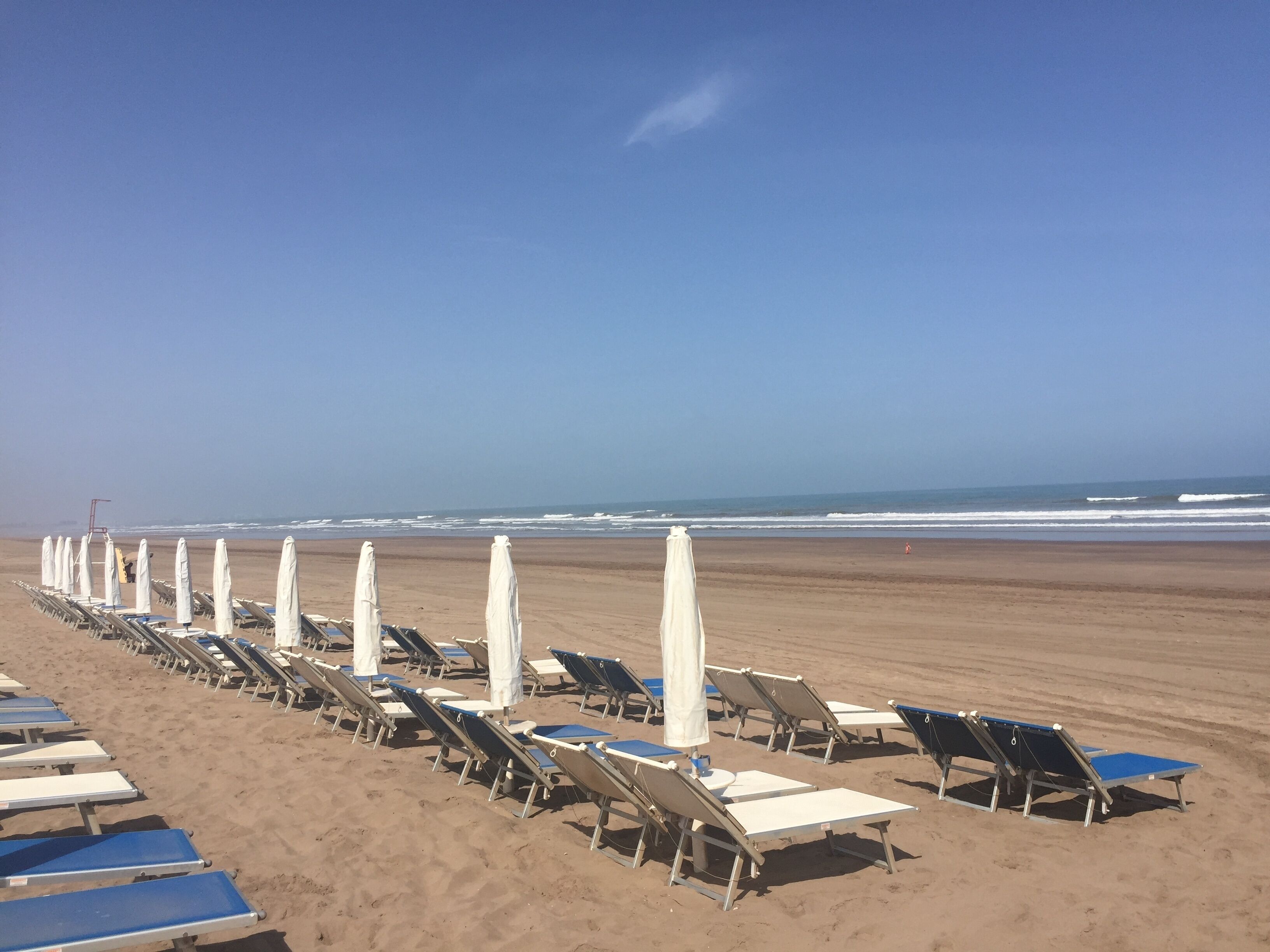 A beach with rows of sun loungers and umbrellas, a clear sky, and the ocean in the background.