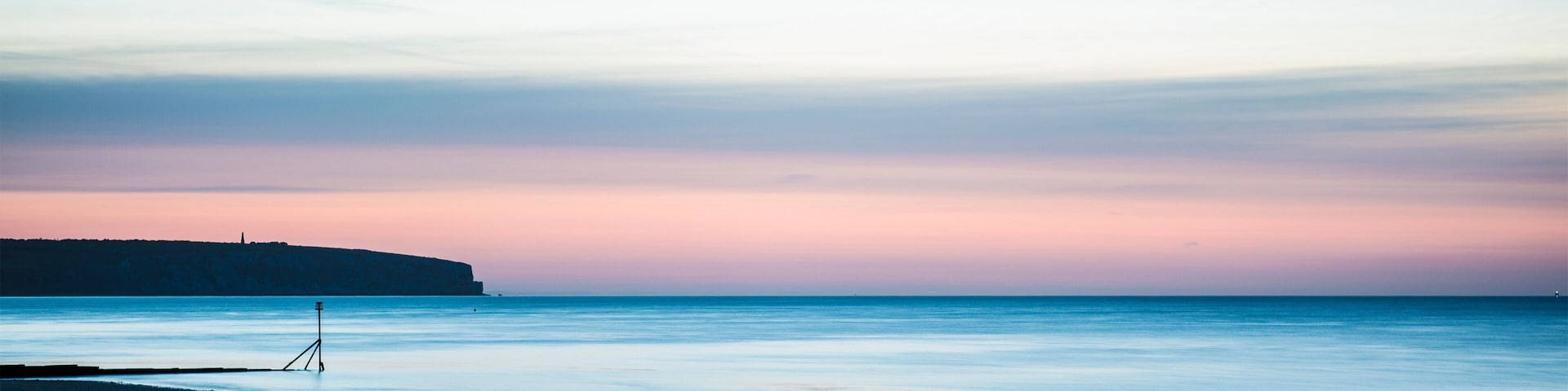 Una playa al atardecer con aguas tranquilas, un muelle y un acantilado distante.