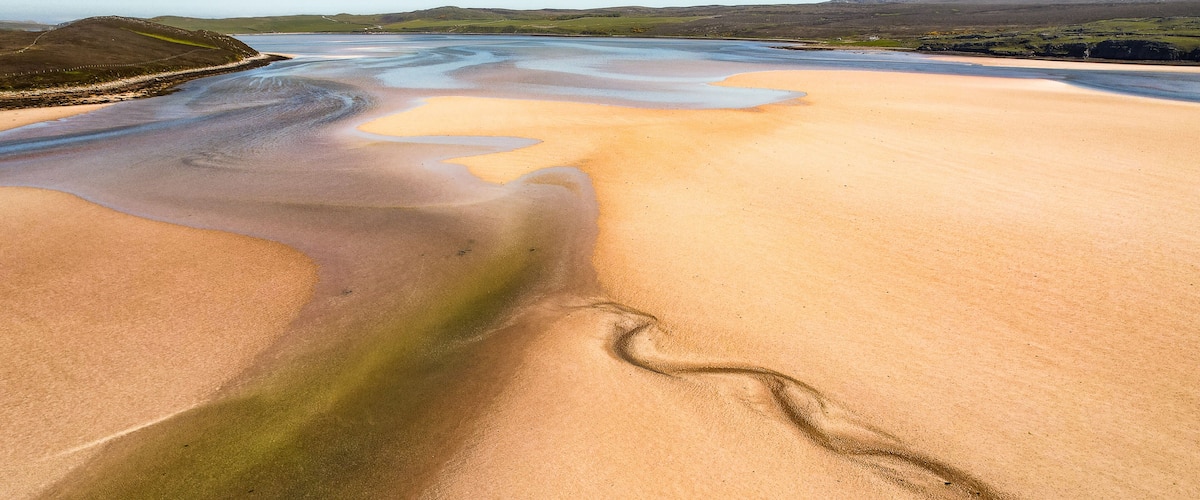 A coastal landscape with a sandy beach, a tidal pool, and distant hills.