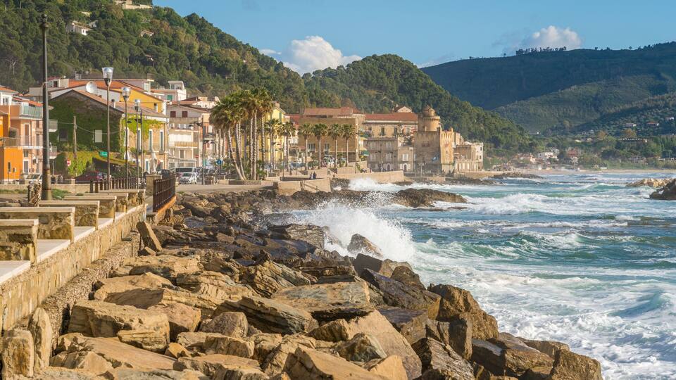 A rocky coastline with waves crashing against the shore, a row of colorful buildings, and a hill with trees in the background.