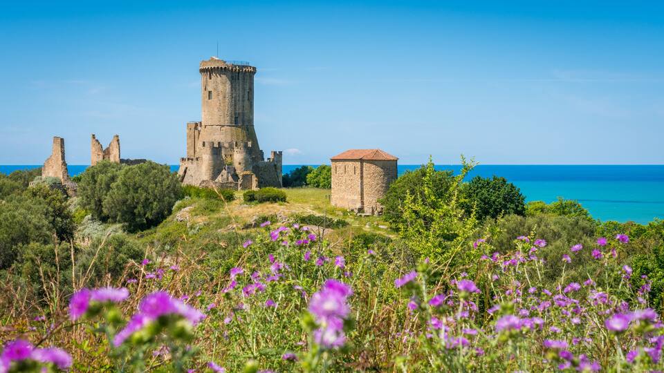 A historic castle with ruins and a tower, surrounded by lush greenery and purple flowers, overlooking a clear blue sea.