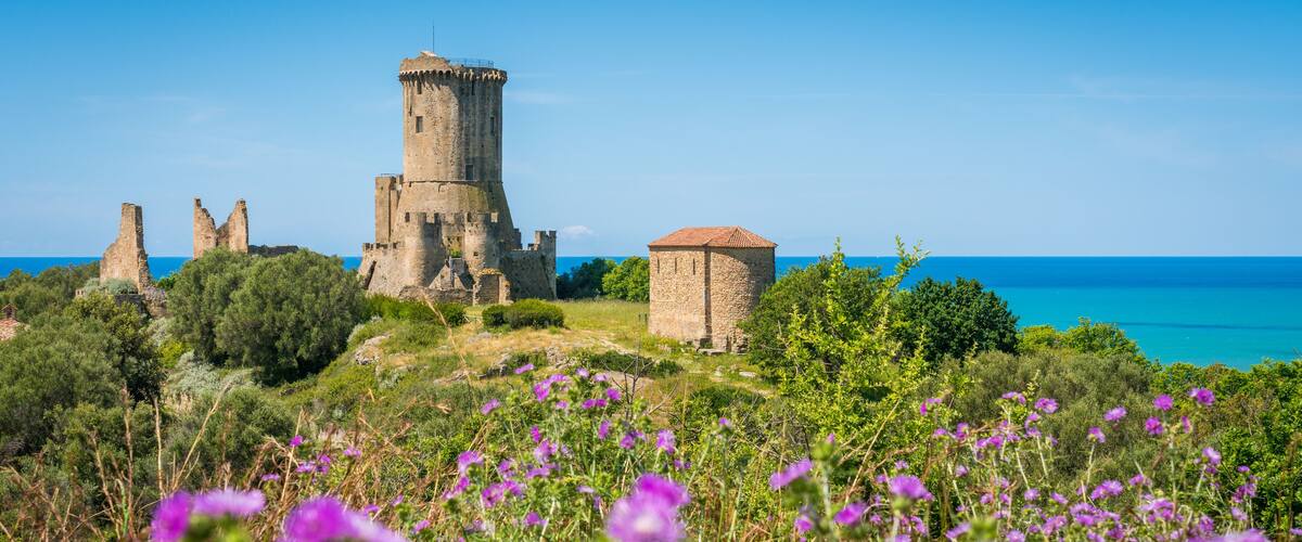 A historic castle with ruins and a tower, surrounded by lush greenery and purple flowers, overlooking a clear blue sea.