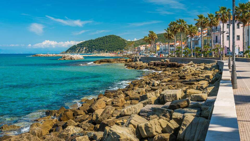 A rocky coastline with clear turquoise waters, a sandy beach, and a row of palm trees and buildings along the shore.
