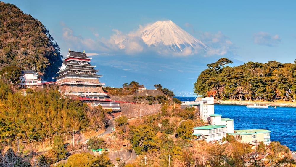 A traditional Japanese castle, Mt. Fuji in the background, and a coastal area with boats.