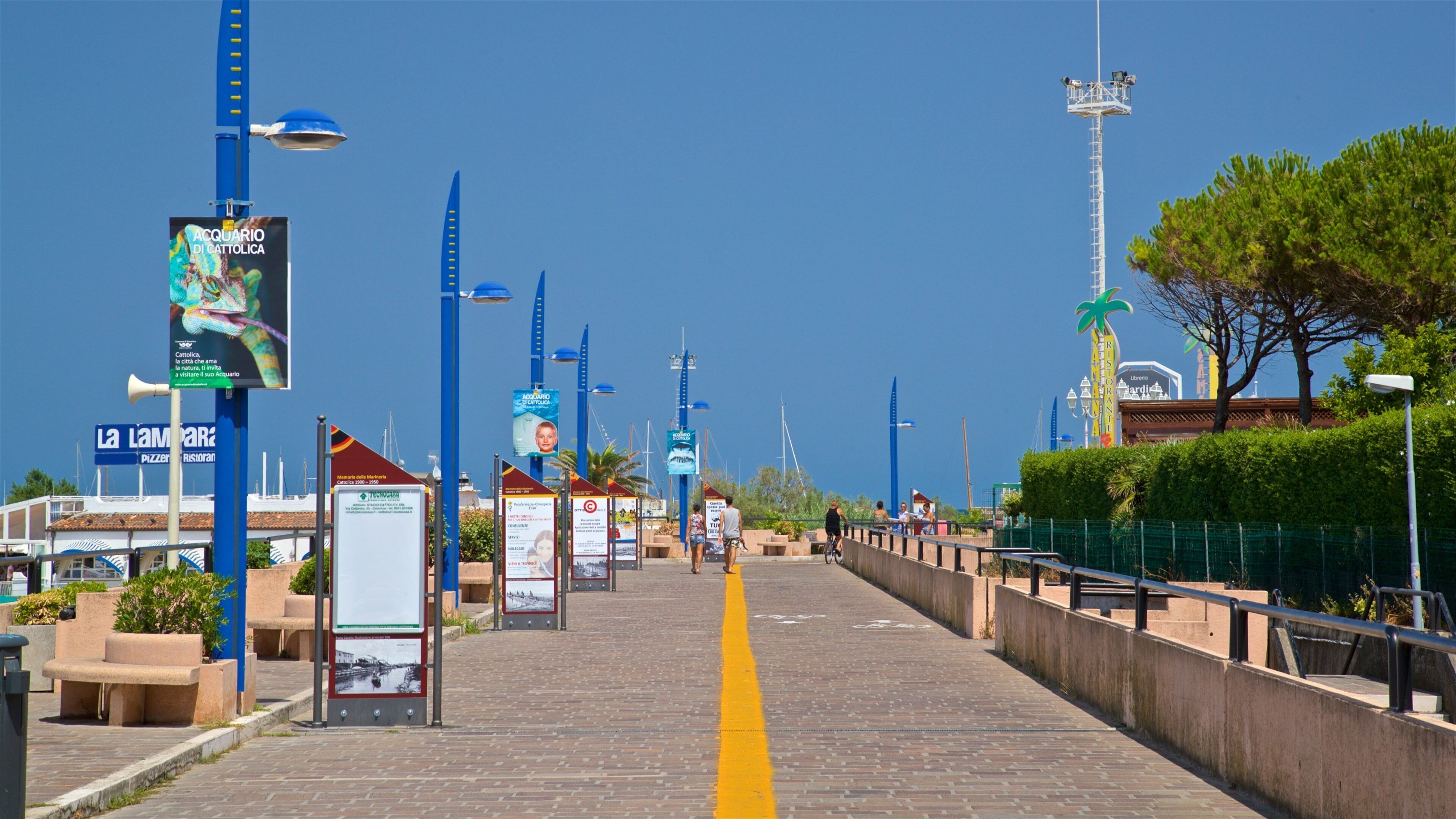 Una passeggiata lungomare con lampioni blu, cartelli informativi e un percorso giallo.