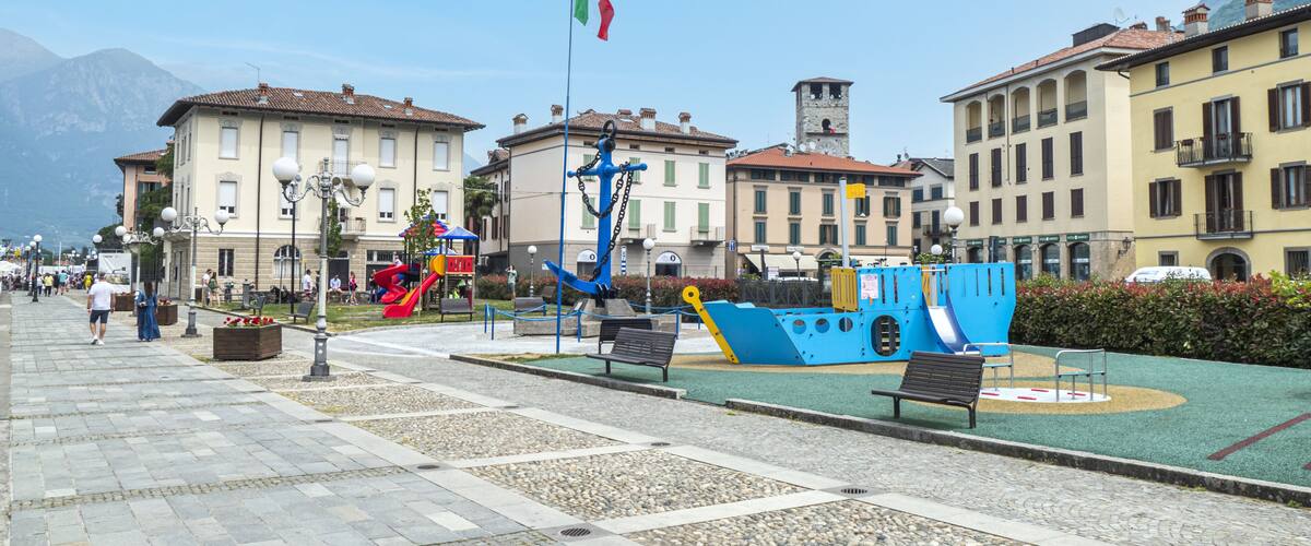 A waterfront promenade with a blue boat sculpture, benches, and a flagpole with the Italian flag.