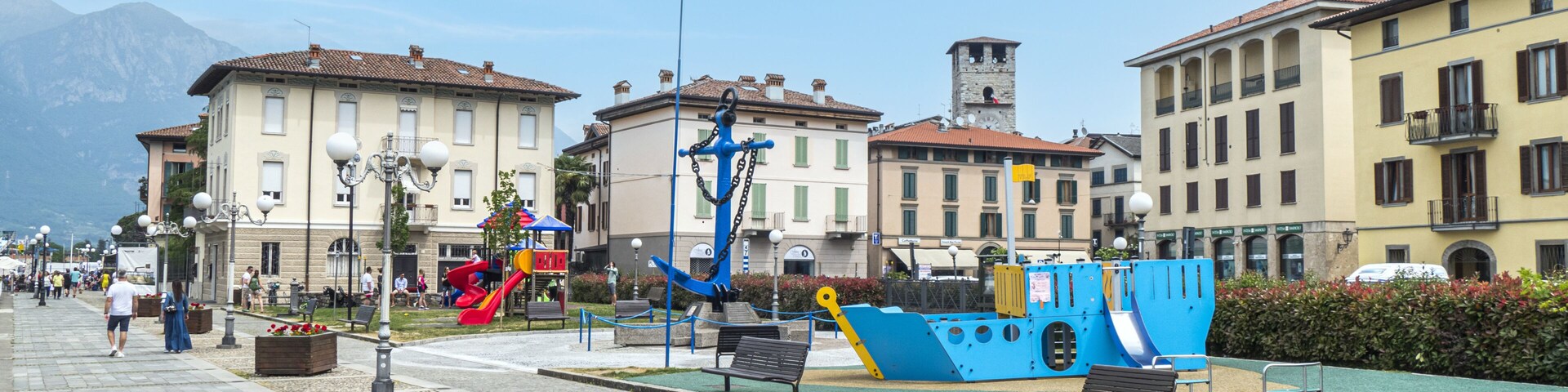 A waterfront promenade with a blue boat sculpture, benches, and a flagpole with the Italian flag.