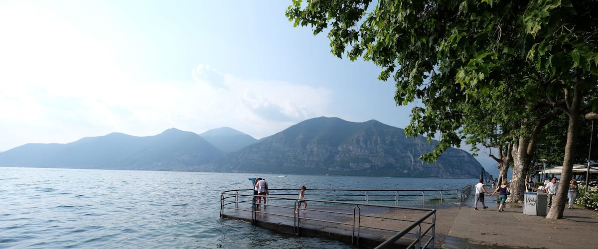 Un paseo a orillas del lago con un muelle que se adentra en el agua, rodeado de montañas y árboles.