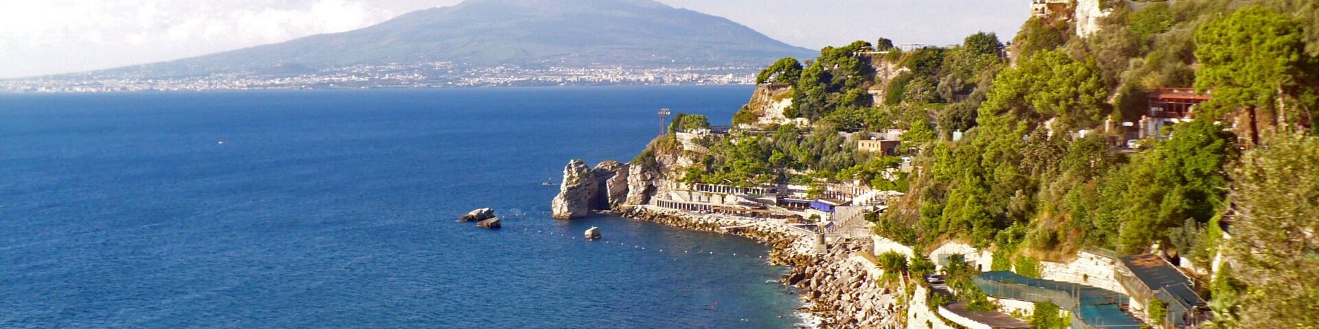 Un pueblo costero con un litoral rocoso, una playa con sombrillas y una montaña al fondo.
