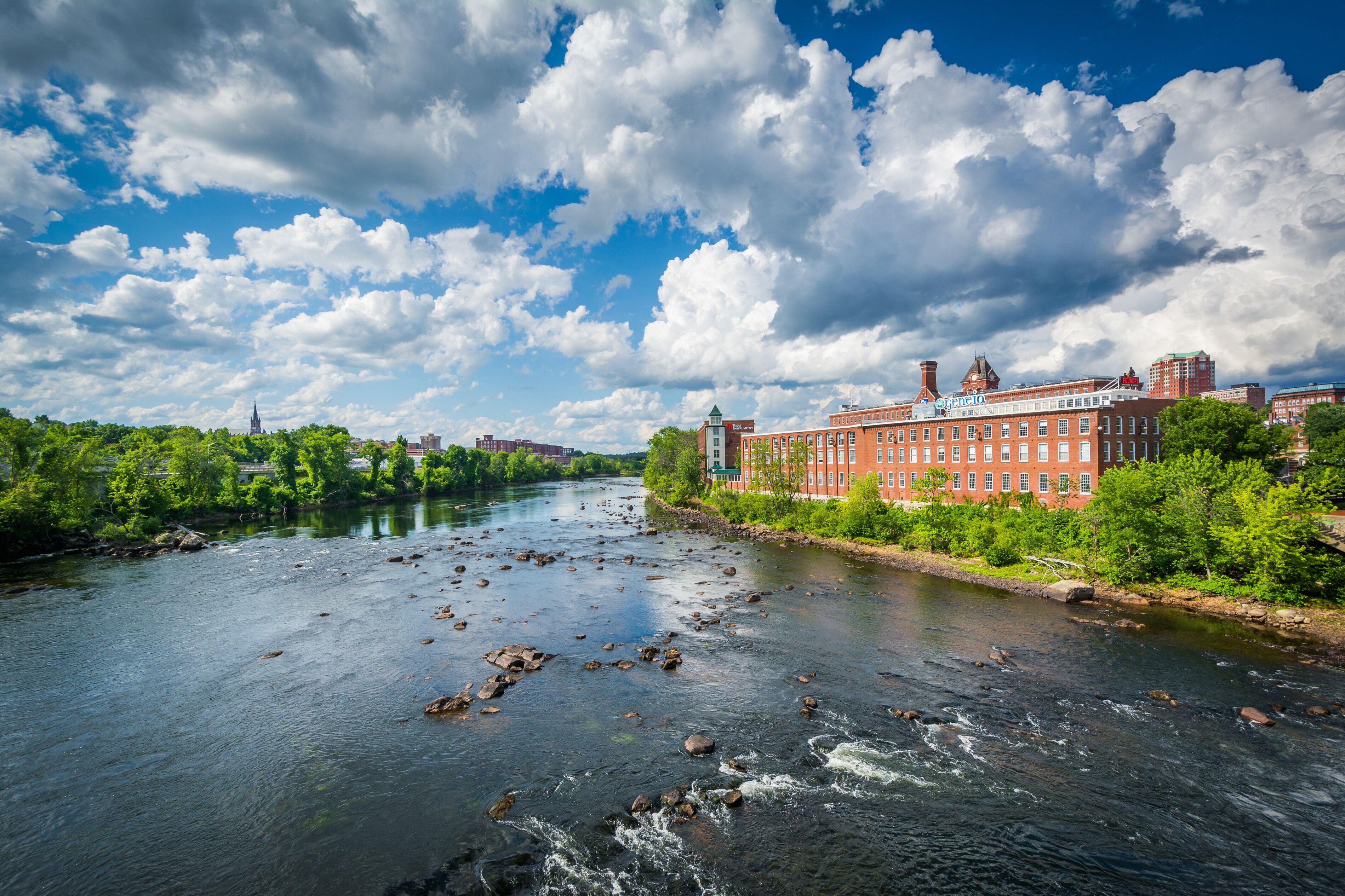 A river with rocks, a red brick building, and green trees under a partly cloudy sky.