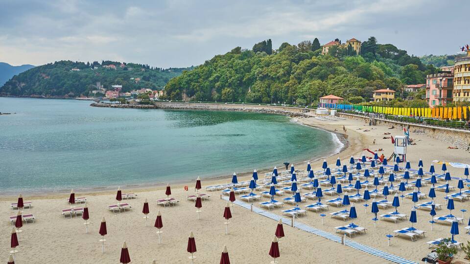 A beach with rows of blue and maroon umbrellas, a sandy shore, and buildings on a hillside in the background.