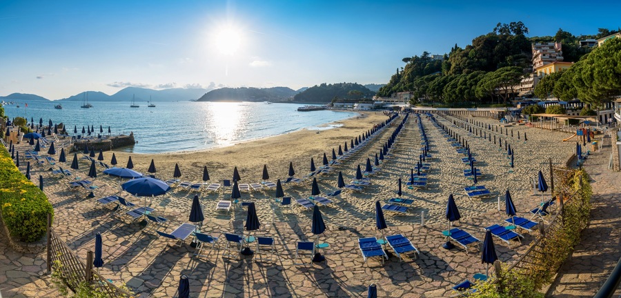 A beach with rows of sun loungers and umbrellas, a clear sky, and boats in the water.