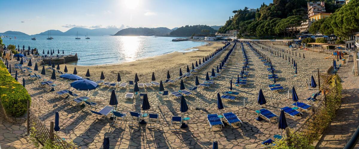 A beach with rows of sun loungers and umbrellas, a clear sky, and boats on the water.