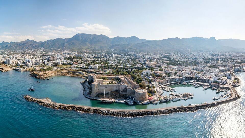 An aerial view of a coastal city with a prominent fortress, a marina, and a mountainous backdrop.