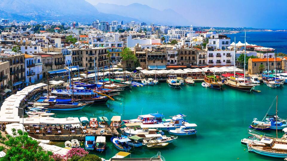 A coastal town with a marina filled with boats, historic buildings, and a mountainous backdrop.