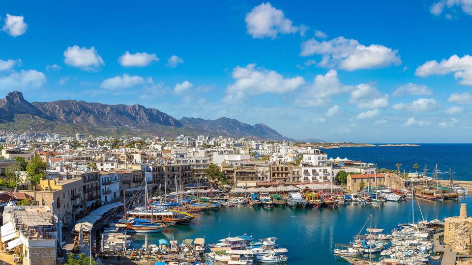 A coastal town with a harbor filled with boats, a lighthouse, and mountains in the background.