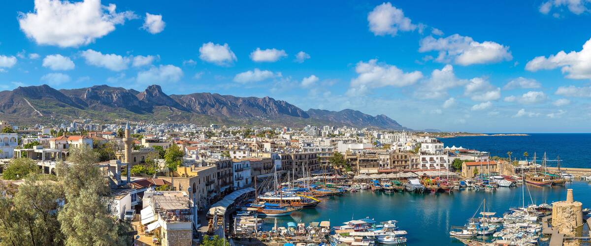A coastal town with a harbor filled with boats, a lighthouse, and mountains in the background.