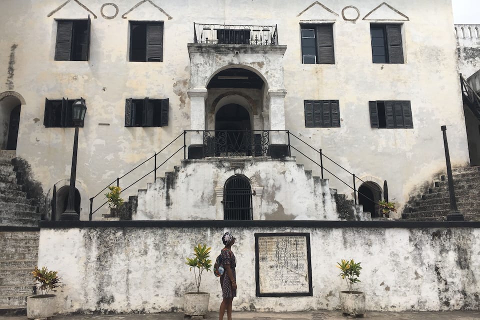 A weathered building with a central archway, multiple windows with shutters, and a staircase leading to a balcony.