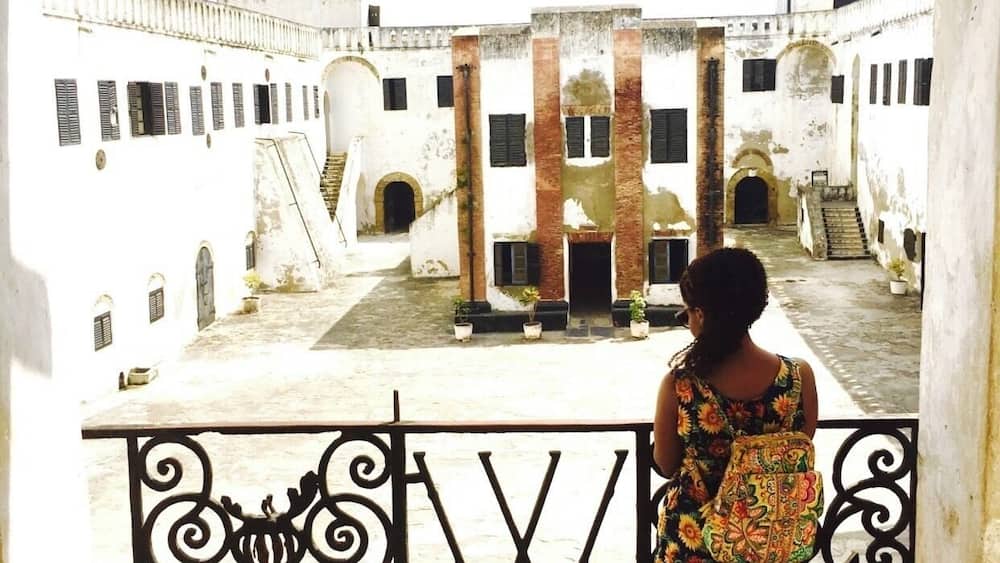 A woman standing on a balcony overlooking a historic courtyard with white buildings and a central staircase.
