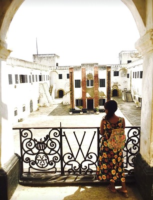 Una mujer de pie en un balcón con vista a un patio histórico con edificios blancos y una escalera central.