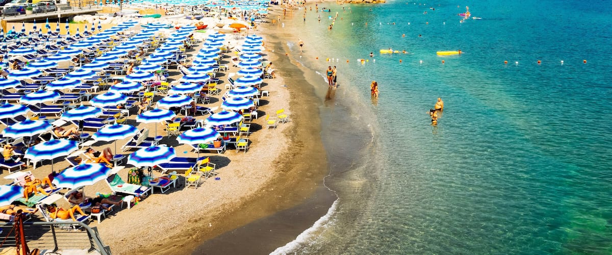 A beach with numerous blue and white umbrellas, people swimming and sunbathing.