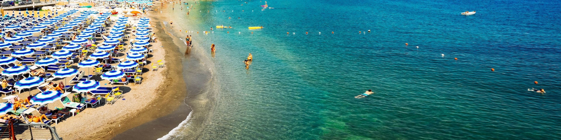 A beach with numerous blue and white umbrellas, people swimming and sunbathing.