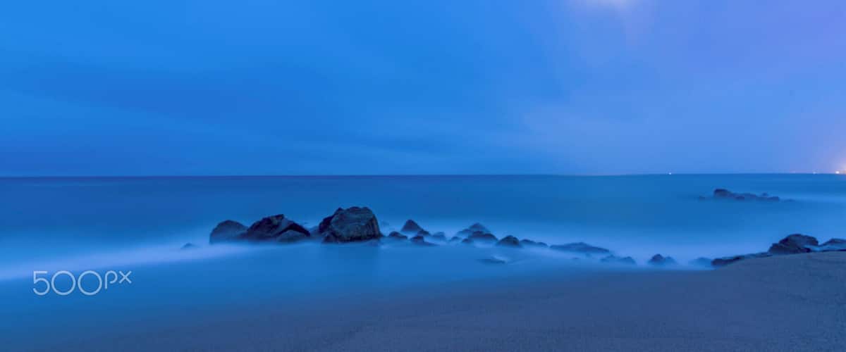 A calm sea at dusk with a visible moon and rocky outcrops along the shore.